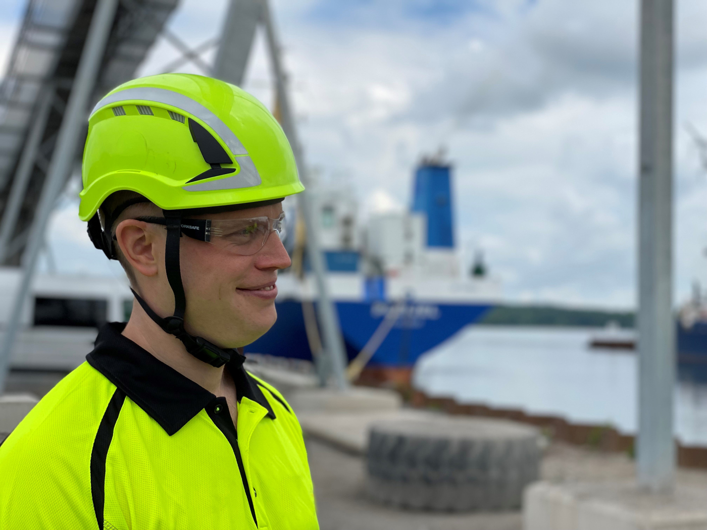 Person i gul skyddshjälm och reflexjacka står nära en hamn med en stor blåvit fraktbåt i bakgrunden, samt en metallstruktur och kajmiljö.
