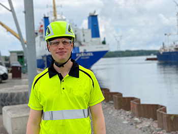 Person klädd i ljusgul skyddströja och hjälm står vid en hamn, med fartyg i bakgrunden.