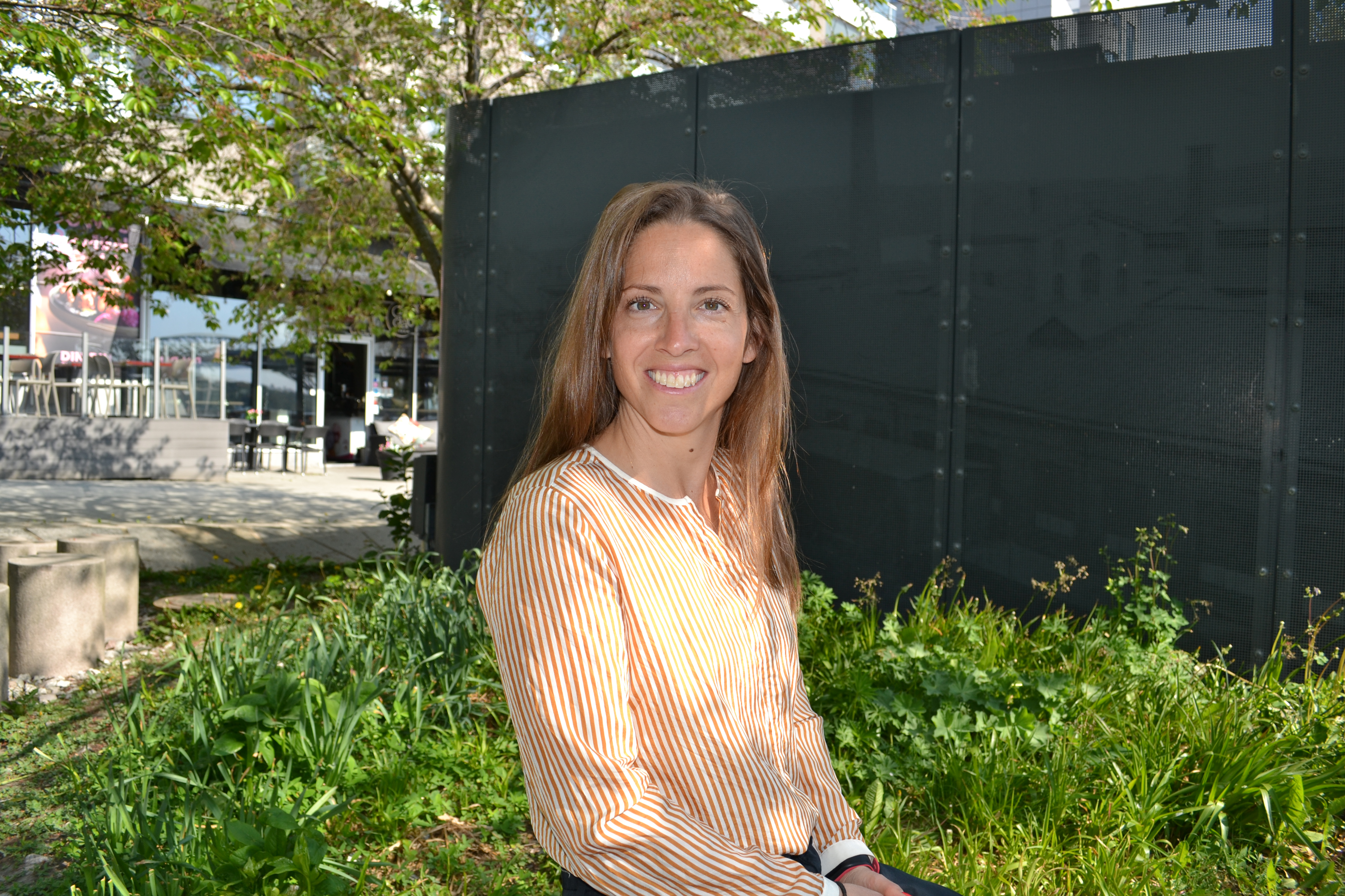 En person sitter på gräs framför en svart vägg, omgiven av grönska och en trädgårdsbakgrund. I bakgrunden syns byggnader och en trädstam i vänstra delen av bilden.