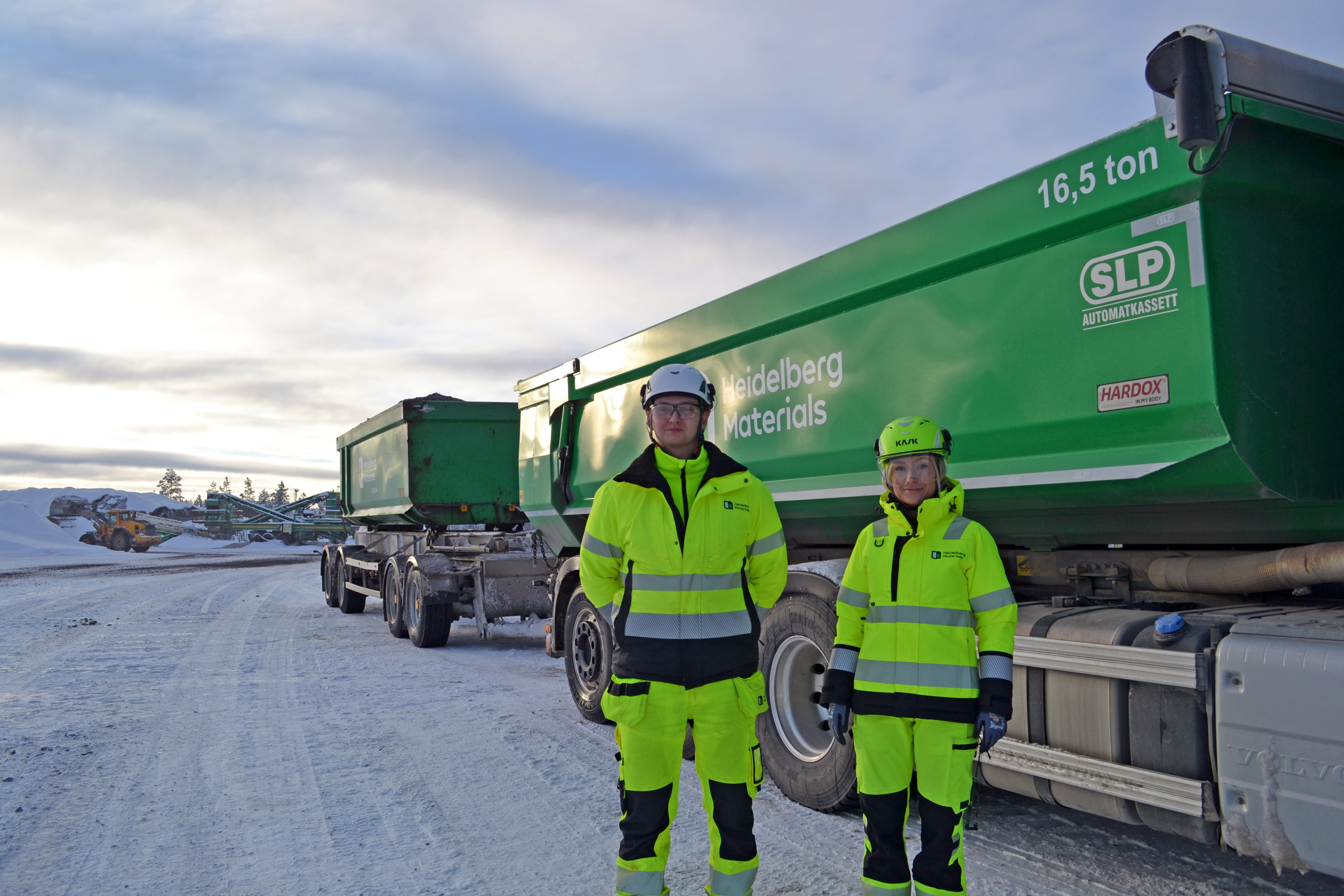 Två personer i gula arbetskläder och hjälmar står framför två gröna lastbilar med tippflak, märkta med 16,5 ton och företagslogotyper. Bakgrunden visar en snöig miljö med molntäckt himmel och maskiner i fjärran.