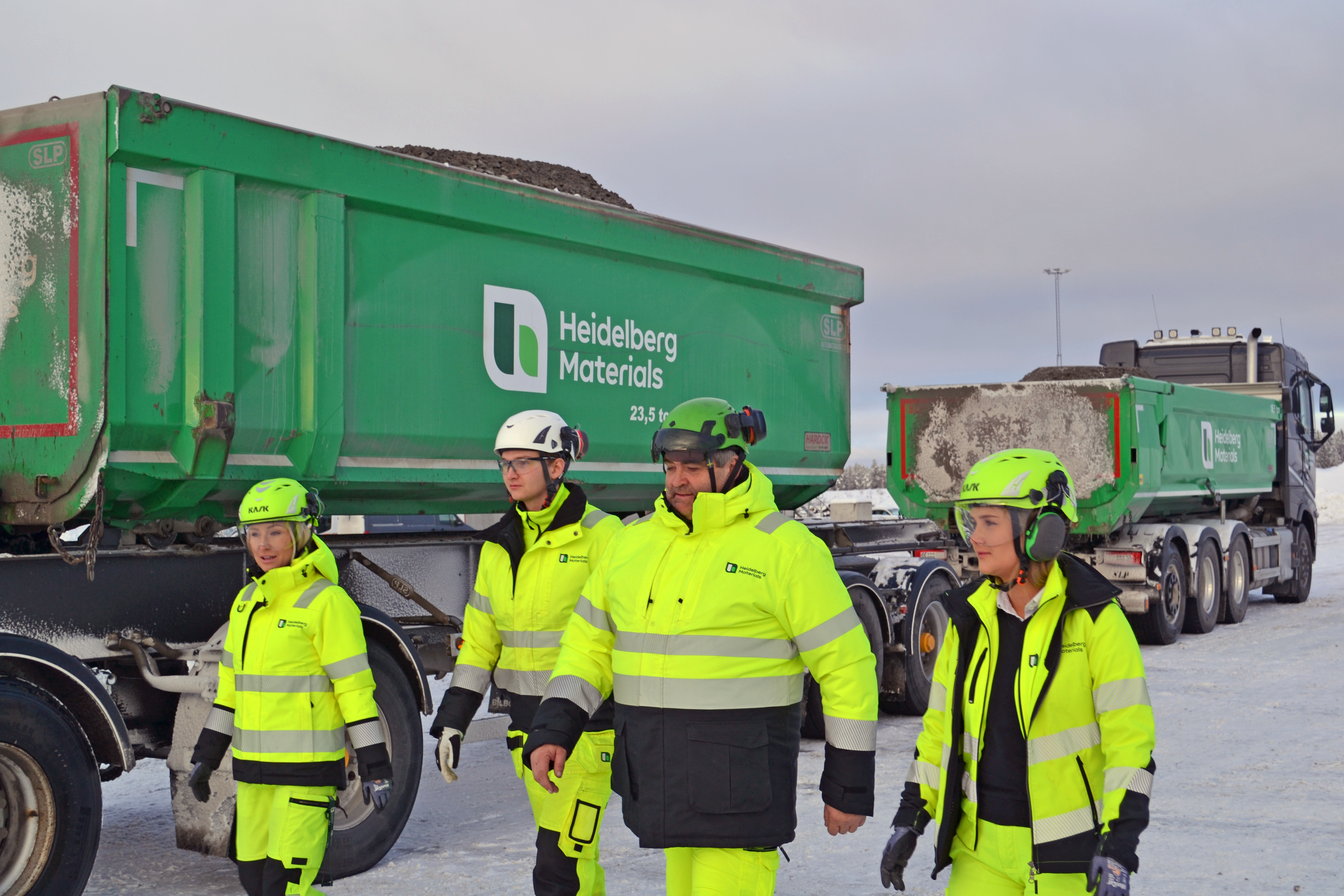 Fyra arbetsklädda personer i gula högsynliga kläder och skyddsutrustning står framför två lastbilar märkta "Heidelberg Materials". Omgivningen är snöig och grå, med fokus på personerna som ser ut att inspektera eller konversera.