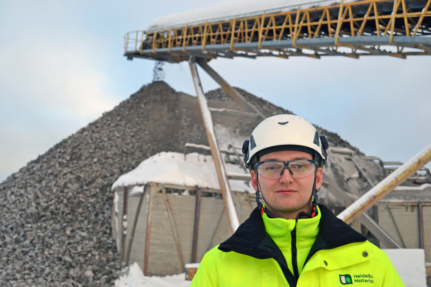 Person i gul säkerhetsjacka och hjälm framför en sten- och grusverksamhet med stora högar av material och ett transportband som sträcker sig diagonalt över bilden. Snö på både marken och utrustningen.