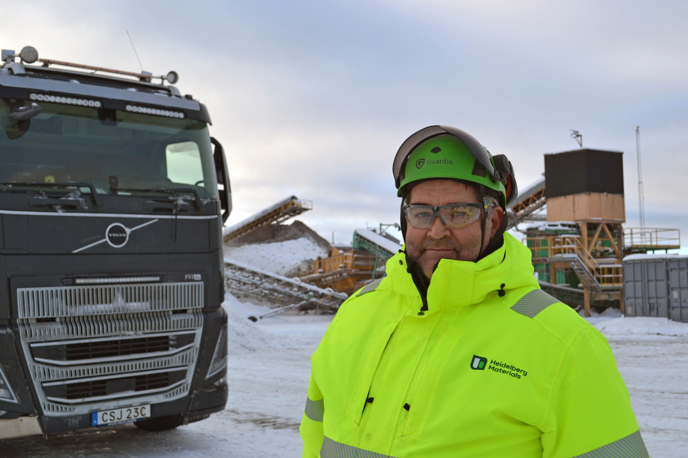 Person klädd i gul varseljacka står bredvid en lastbil på en snötäckt arbetsplats med byggmaterial i bakgrunden.