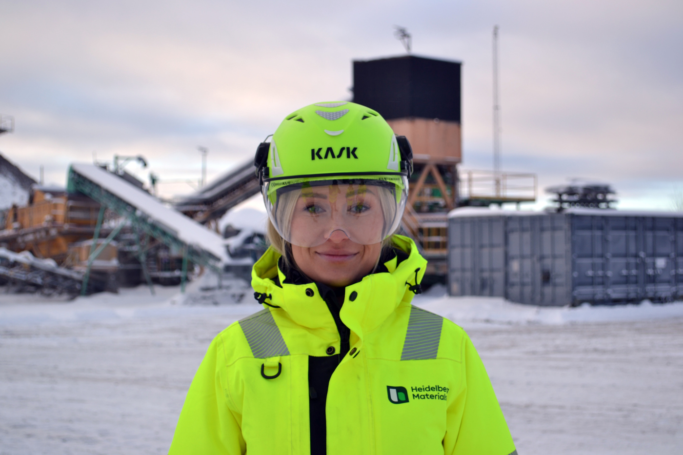 En person iklädd gul skyddsutrustning med hjälm står på en industriell arbetsplats i en snöig utomhusmiljö. I bakgrunden syns utrustning som transportband, containrar och byggnader.