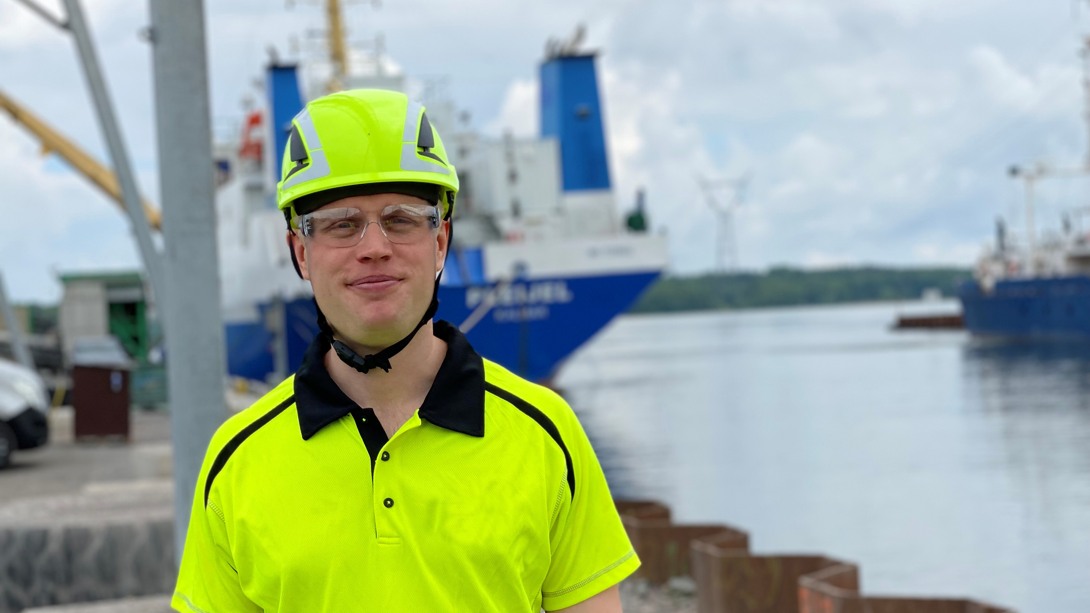 Person i neongul skyddshjälm och reflextröja står vid en hamn. I bakgrunden syns fartyg, vatten och kranar under en molnig himmel.