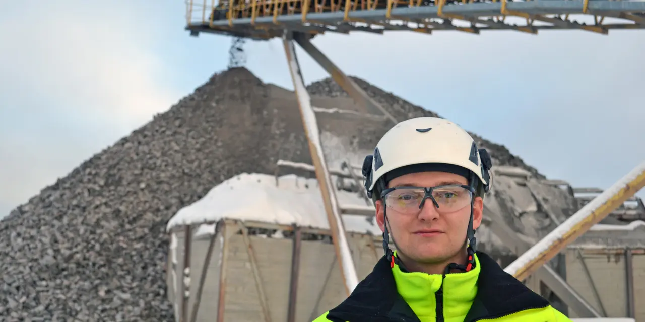 Person i gul säkerhetsjacka och hjälm framför en sten- och grusverksamhet med stora högar av material och ett transportband som sträcker sig diagonalt över bilden. Snö på både marken och utrustningen.
