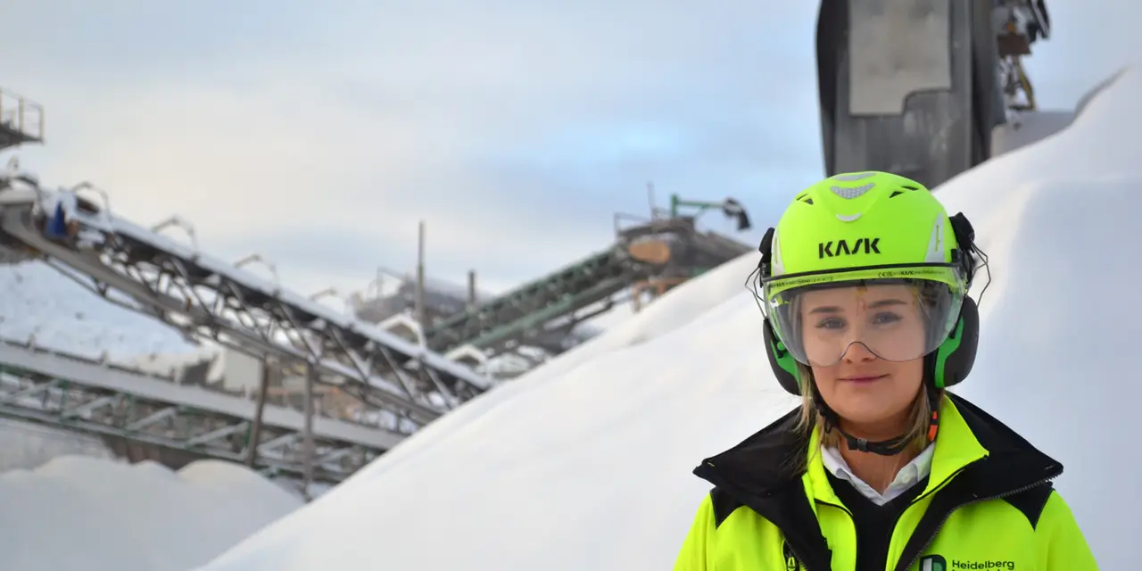 Person i gul skyddsjacka och hjälm står framför en industriell miljö med transportband och stora vita högar av material, sannolikt sand eller snö, under en mulen himmel.
