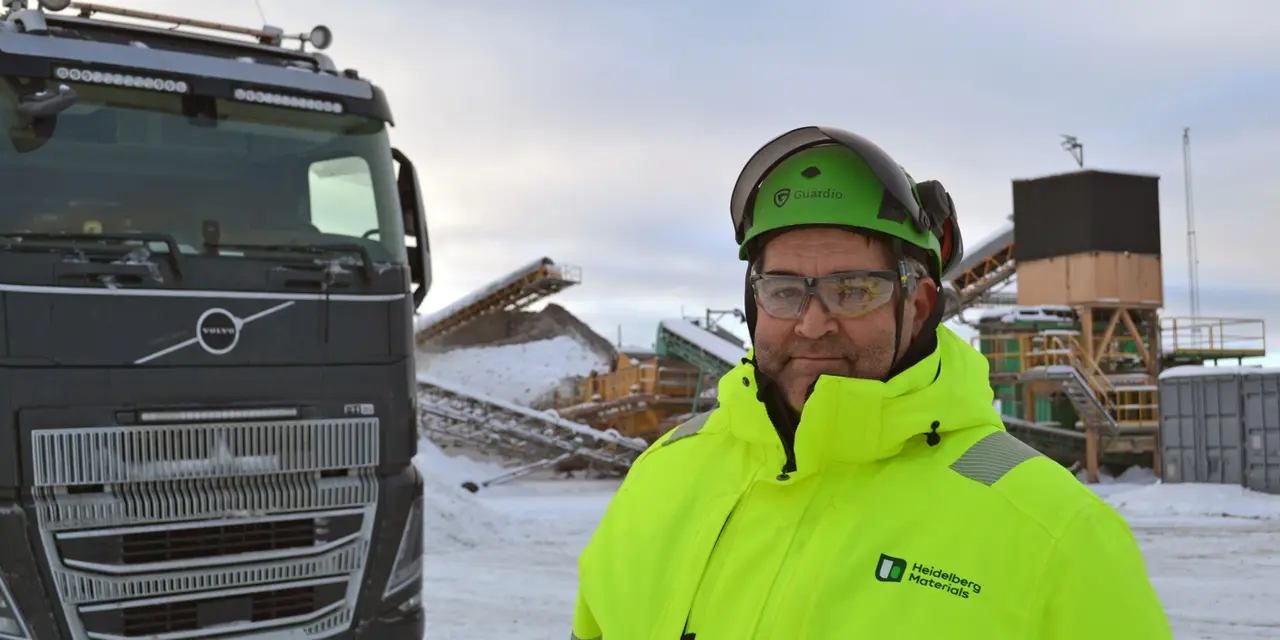 Person klädd i gul varseljacka står bredvid en lastbil på en snötäckt arbetsplats med byggmaterial i bakgrunden.