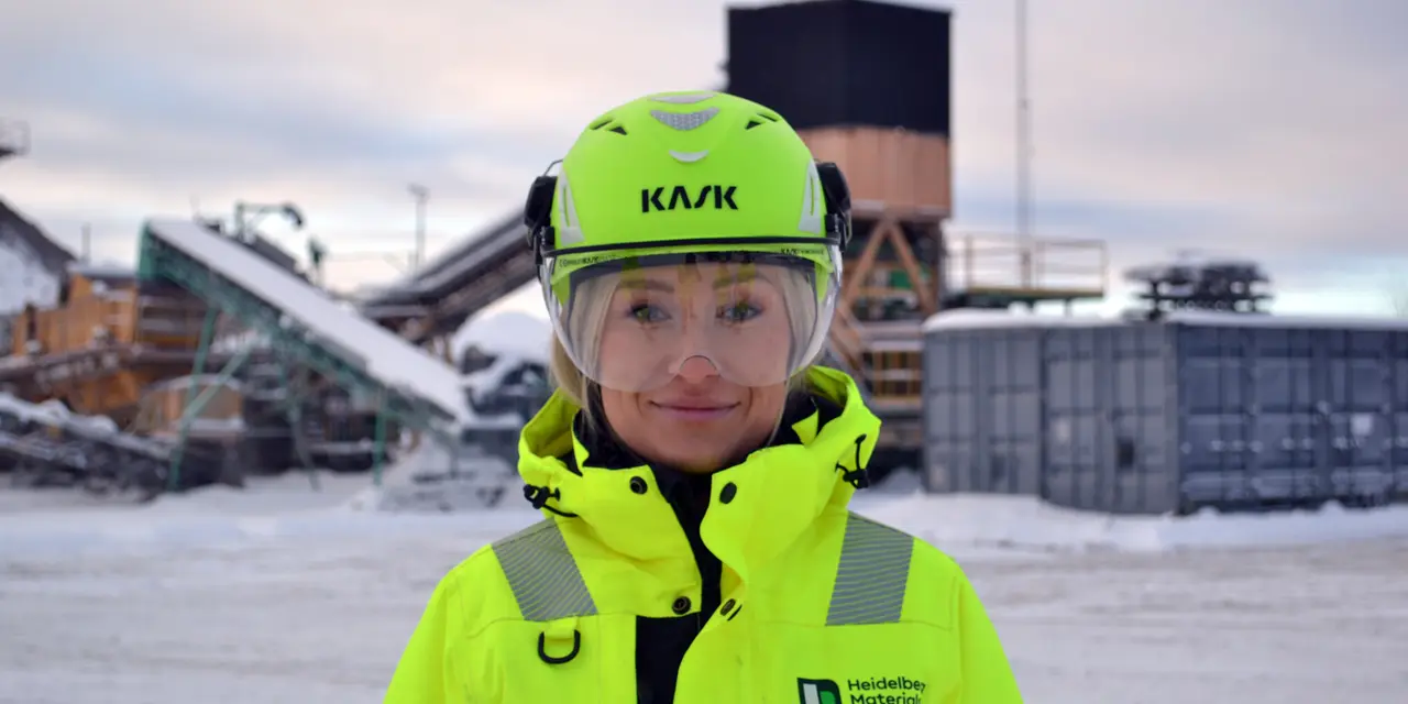 En person iklädd gul skyddsutrustning med hjälm står på en industriell arbetsplats i en snöig utomhusmiljö. I bakgrunden syns utrustning som transportband, containrar och byggnader.