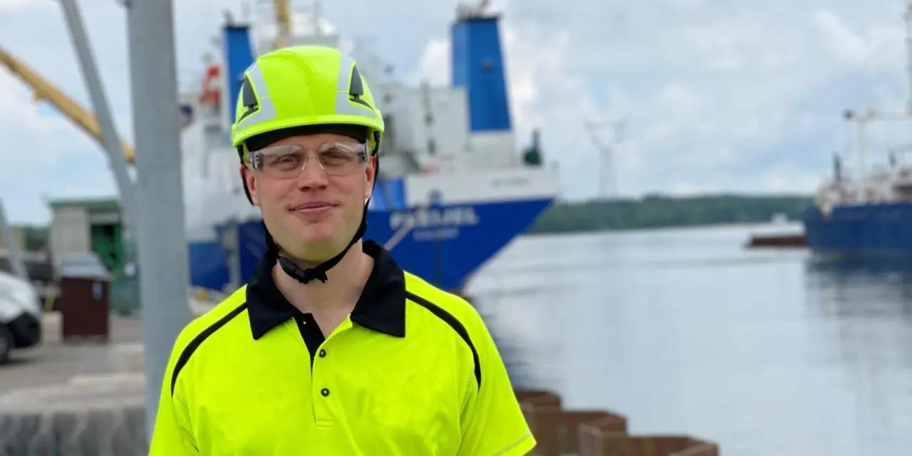 Person i neongul skyddshjälm och reflextröja står vid en hamn. I bakgrunden syns fartyg, vatten och kranar under en molnig himmel.