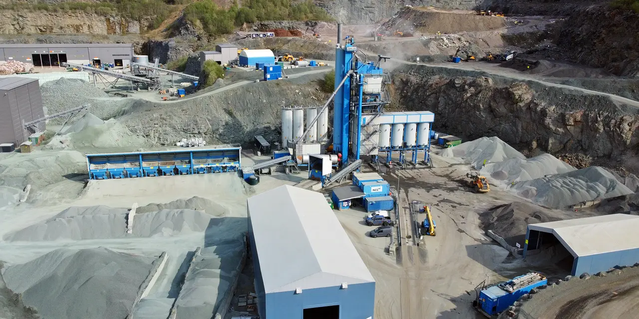 A large industrial Asphalt site with blue and white equipment for processing building materials, surrounded by piles of gravel and storage buildings.