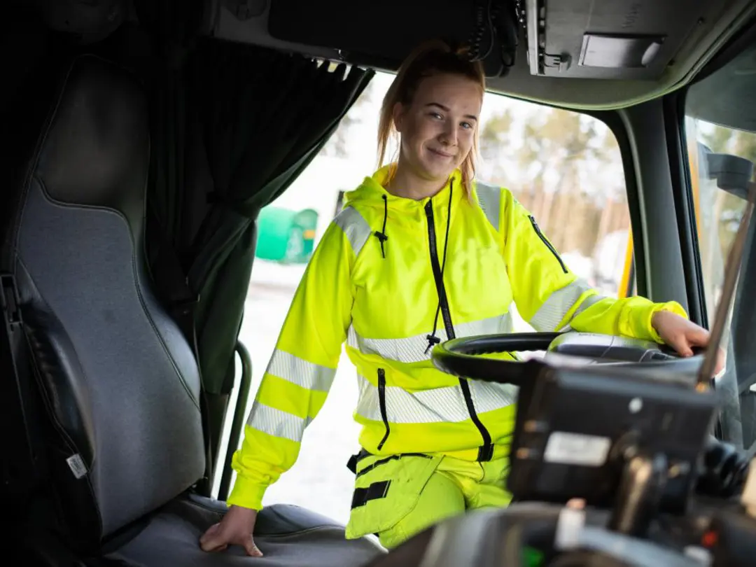 Person i neongula arbetskläder står vid förarplatsen i en lastbil, med ena handen på ratten och den andra vilande på sätet. Bakgrunden visar ett utomhusområde med träd och suddiga detaljer.