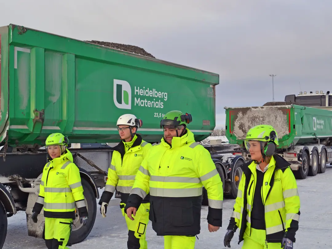 Fyra arbetsklädda personer i gula högsynliga kläder och skyddsutrustning står framför två lastbilar märkta "Heidelberg Materials". Omgivningen är snöig och grå, med fokus på personerna som ser ut att inspektera eller konversera.