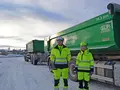 Två personer i gula arbetskläder och hjälmar står framför två gröna lastbilar med tippflak, märkta med 16,5 ton och företagslogotyper. Bakgrunden visar en snöig miljö med molntäckt himmel och maskiner i fjärran.