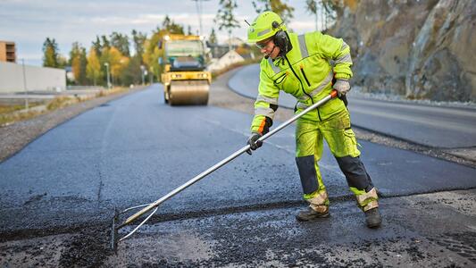 A construction worker in high-visibility clothing smooths fresh asphalt on a road, with a steamroller visible in the background.