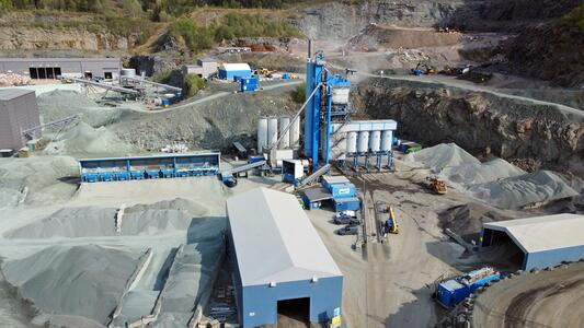 A large industrial Asphalt site with blue and white equipment for processing building materials, surrounded by piles of gravel and storage buildings.